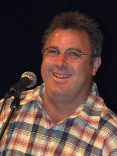 a man with glasses is smiling while holding a microphone in front of him and wearing a plaid shirt