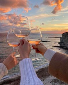 two people toasting wine glasses with the ocean and sky in the background at sunset