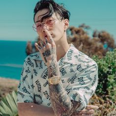 a man with tattoos on his arms and hands is standing near the ocean while holding his hand up to his face