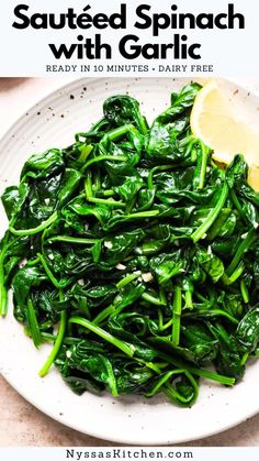 a white plate topped with spinach covered in garlic and lemon wedges next to a slice of lemon