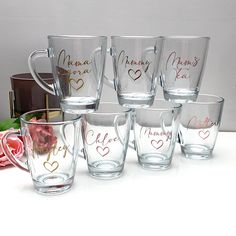 six personalized glass mugs with names and hearts on the side, sitting next to a pink rose