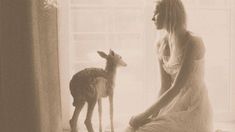 a woman in white dress sitting next to a baby deer on the floor near a window