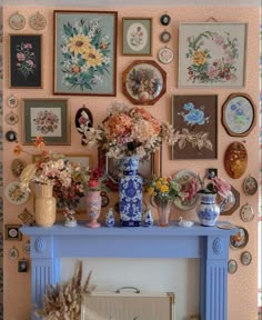 a blue mantle topped with lots of vases filled with flowers next to a wall covered in pictures