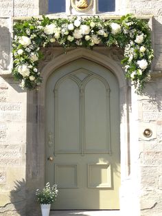 a green door with white flowers and greenery on the top, in front of a stone building