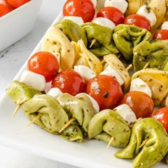 several skewered vegetables are on a white platter with tomatoes and peppers in the background