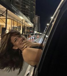 a woman leaning out the window of a car on a city street at night with buildings in the background