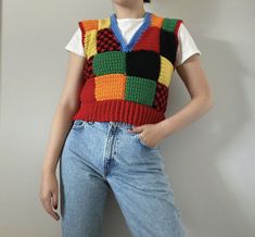 a woman wearing a multicolored knitted sweater vest and jeans stands in front of a white wall