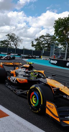 an orange race car driving down a track with other cars behind it on a cloudy day