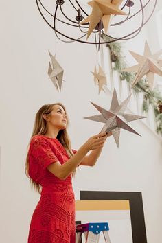 a woman in a red dress is holding up some paper stars that are hanging from a chandelier