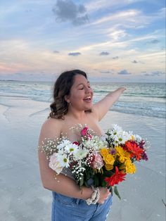 a woman on the beach with flowers in her hand and pointing to the sky above her