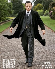 a man in a suit and tie walking down a gravel road with trees behind him