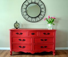 a red dresser with flowers and a clock on the wall next to it in front of a round mirror