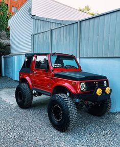 a red and black jeep parked in front of a blue wall with large tires on it