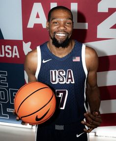 a man holding a basketball in his right hand and smiling at the camera while standing next to a wall