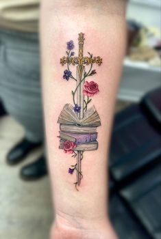 a cross and book tattoo on the left inner arm, with flowers growing out of it