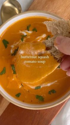 a hand holding a piece of bread over a bowl of butternut squash tomato soup