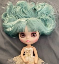 a doll with green hair is sitting on a gray sheet and wearing a white dress