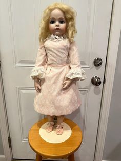 a doll sitting on top of a wooden stool in front of a white door with a pink dress