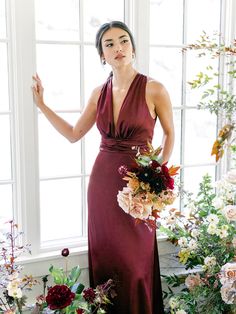 a woman standing in front of a window wearing a long dress and holding a bouquet