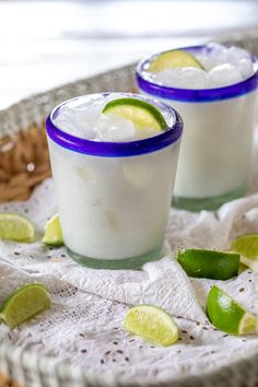 two glasses filled with ice and limes on top of a white cloth covered tray