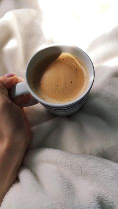 a person holding a cup of coffee on top of a white bed sheet with their hands