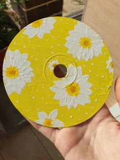 a hand holding a yellow and white painted disc with daisies on it, in front of a plant