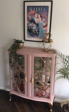 a pink cabinet with glass doors and gold handles in front of a painting on the wall