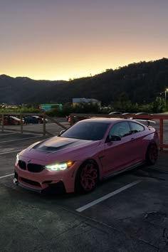 a pink car parked in a parking lot next to a fence and mountains at sunset