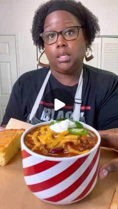 a woman holding a bowl of chili with cheese and bread in front of her face