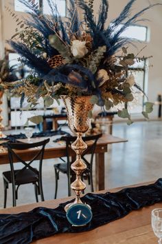 a tall vase filled with blue flowers and greenery on top of a wooden table