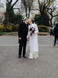 a bride and groom kissing in the middle of a park with people walking around behind them