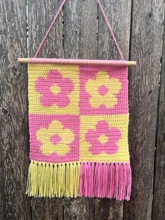 a pink and yellow crocheted wall hanging on a wooden fence with fringes