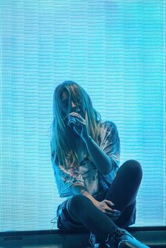 a man with long hair sitting on the ground in front of a large screen holding a microphone