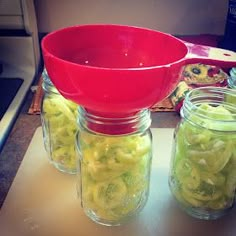 four jars filled with pickles on top of a counter next to a red strainer