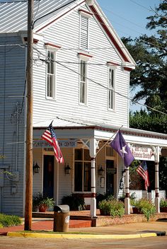 an old white building with two american flags on the front and one purple flag on the back