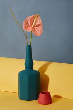 a blue vase with a pink flower in it next to a red cup on a yellow table