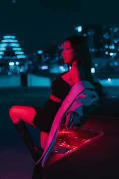 a woman is sitting on the hood of a car in front of a city at night