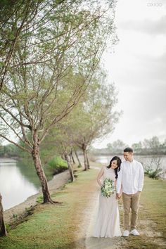 a man and woman standing next to each other on a dirt road near water with trees in the background