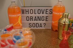 an orange soda sign sitting on top of a counter next to other drinks and condiments