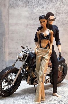 a barbie doll is standing next to a motorcycle with a man on the bike behind it