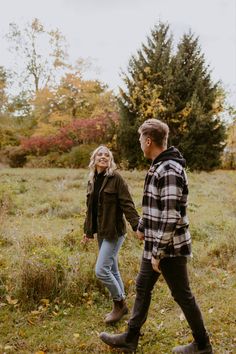 a man and woman holding hands walking through the grass in an open field with trees behind them