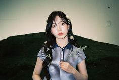 a woman with long black hair standing in front of a green hill holding a flower