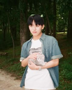 a young man holding a cat in his hands while standing on a dirt road surrounded by trees