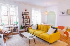 a living room with yellow couches and white curtains on the windowsill, bookshelf in the background