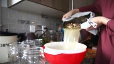 a person pouring something into a red bowl on the kitchen counter with other jars and containers