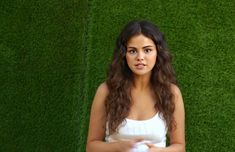 a woman standing in front of a green grass wall with her hands together and looking at the camera