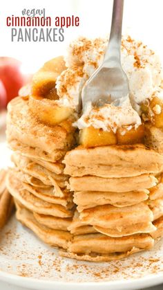 a stack of pancakes topped with whipped cream and apple slices on a white plate next to cinnamon sticks