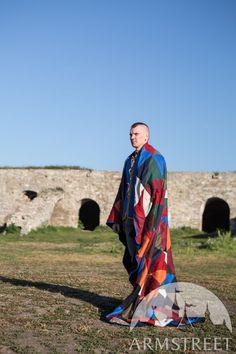 a man standing in the grass with a colorful blanket on his back and an old stone wall behind him