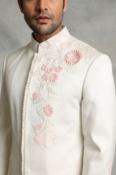 a man in a white suit with pink flowers on the lapel and collar is looking at the camera