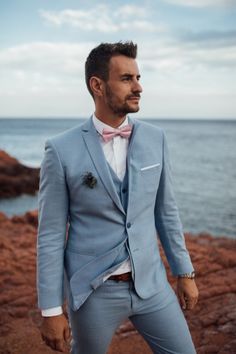 a man in a blue suit and bow tie standing on rocks near the ocean with his hands in his pockets